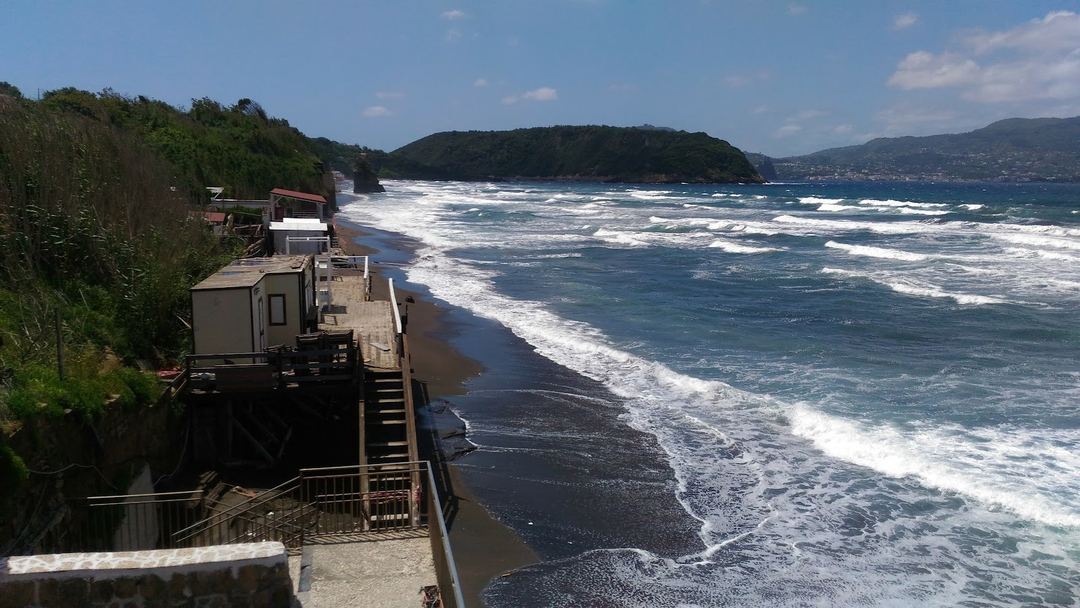 Dramatic coastal view from the property featuring crashing waves, black sand beach, and forested headlands under clear sky.