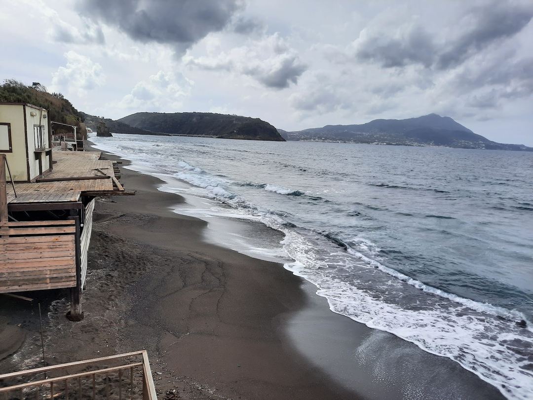 A black sand beach with wooden beach cabins overlooks the turquoise sea with mountains visible across the bay under a dramatic cloudy sky.