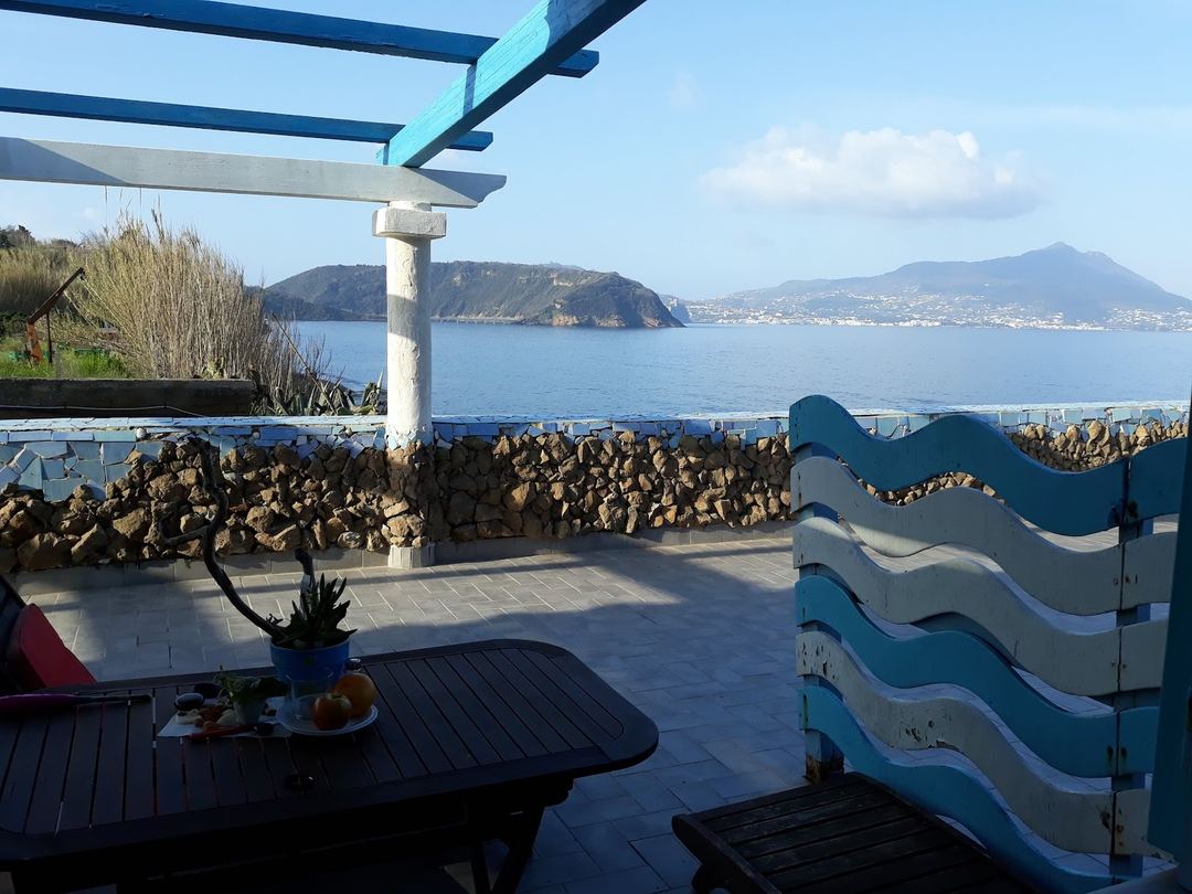 Panoramic coastal view from a terrace overlooking a Mediterranean bay with rocky breakwater and distant mountains under clear skies.